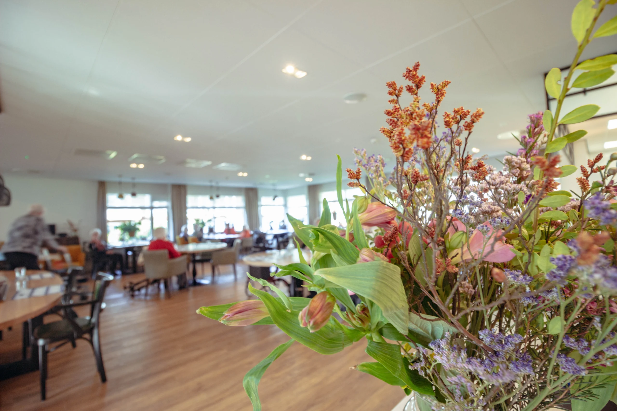 Lichte en ruime gemeenschapsruimte met vaas bloemen op tafel, mensen aan eettafels en houten vloer