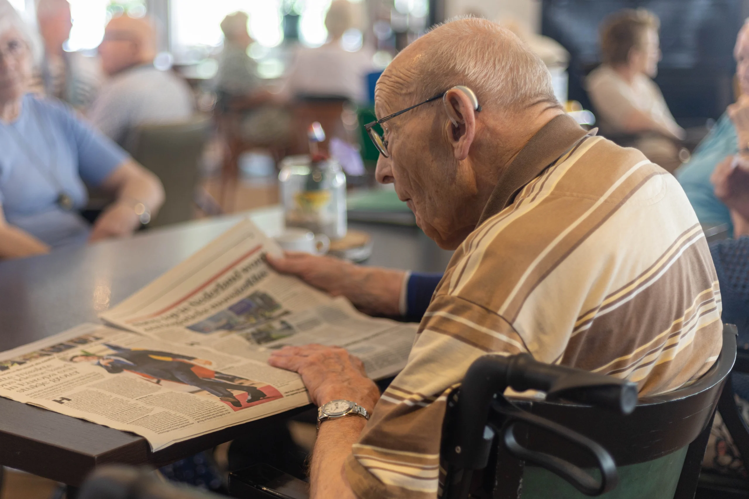 Ouder man in rolstoel leest krant aan tafel met mensen op de achtergrond in lichte ruimte