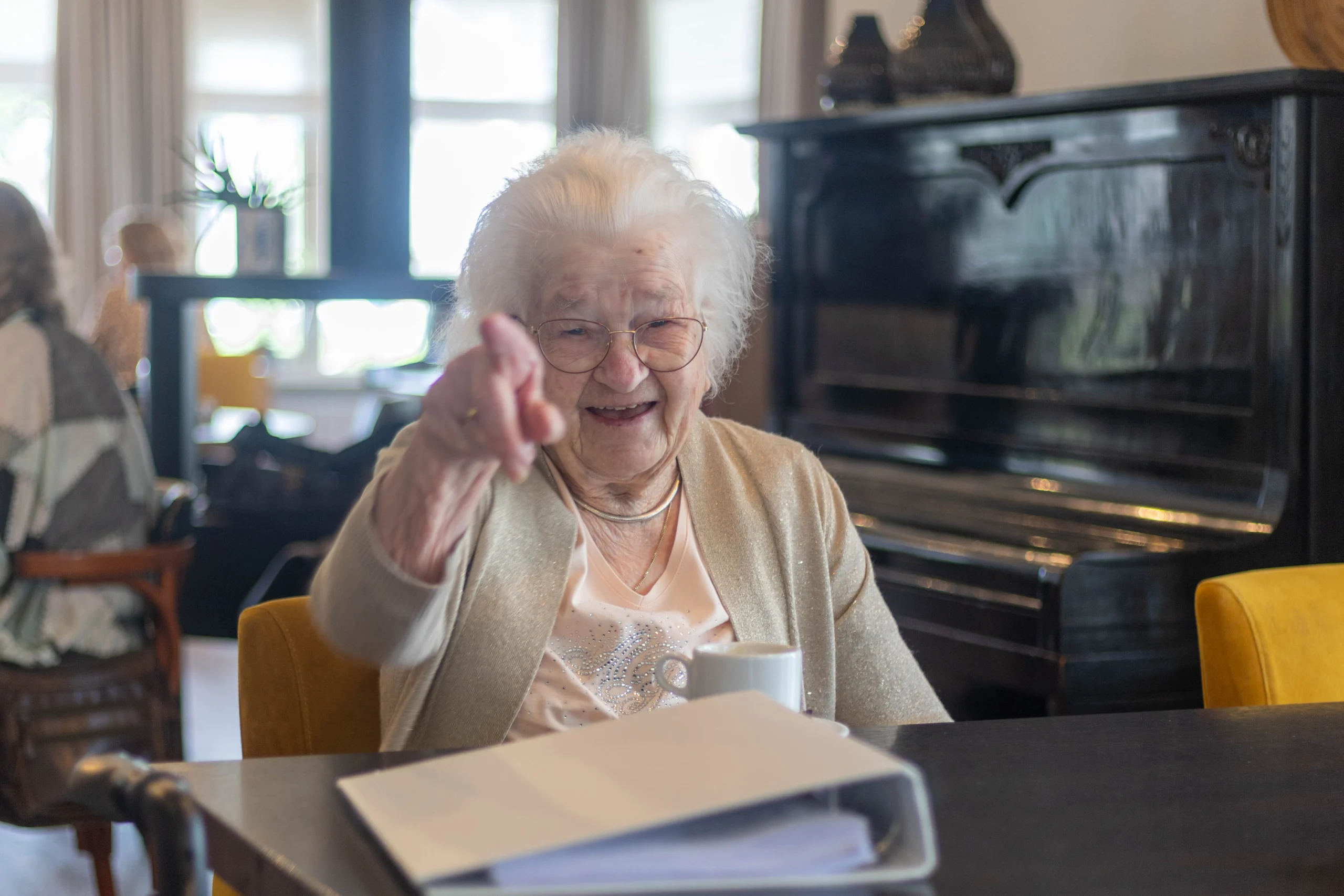 Oudere vrouw met wit haar lacht en wijst zittend aan tafel met map en koffie in een sociale ruimte