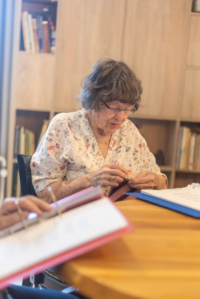 Oudere vrouw breit aan een ronde houten tafel met boekenplanken op de achtergrond