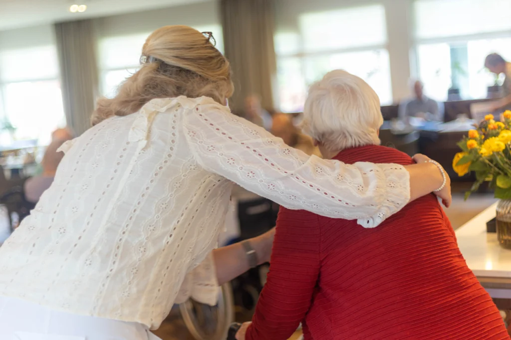 Twee vrouwen in witte blouse en rode trui knuffelen in een lichte ruimte met bloemen op tafel