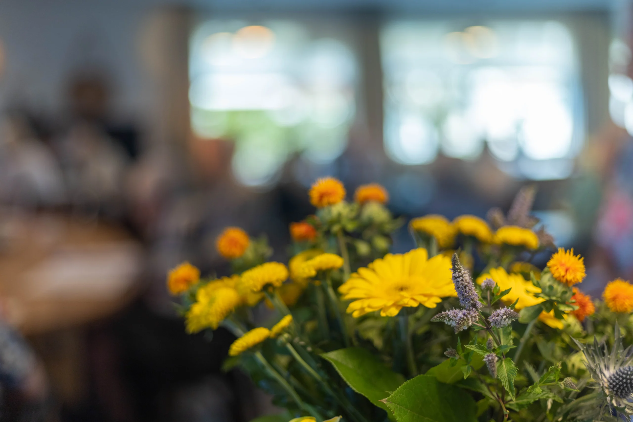 Bloemrijk boeket met heldere gele en oranje bloemen voor een wazige achtergrond met mensen en tafel