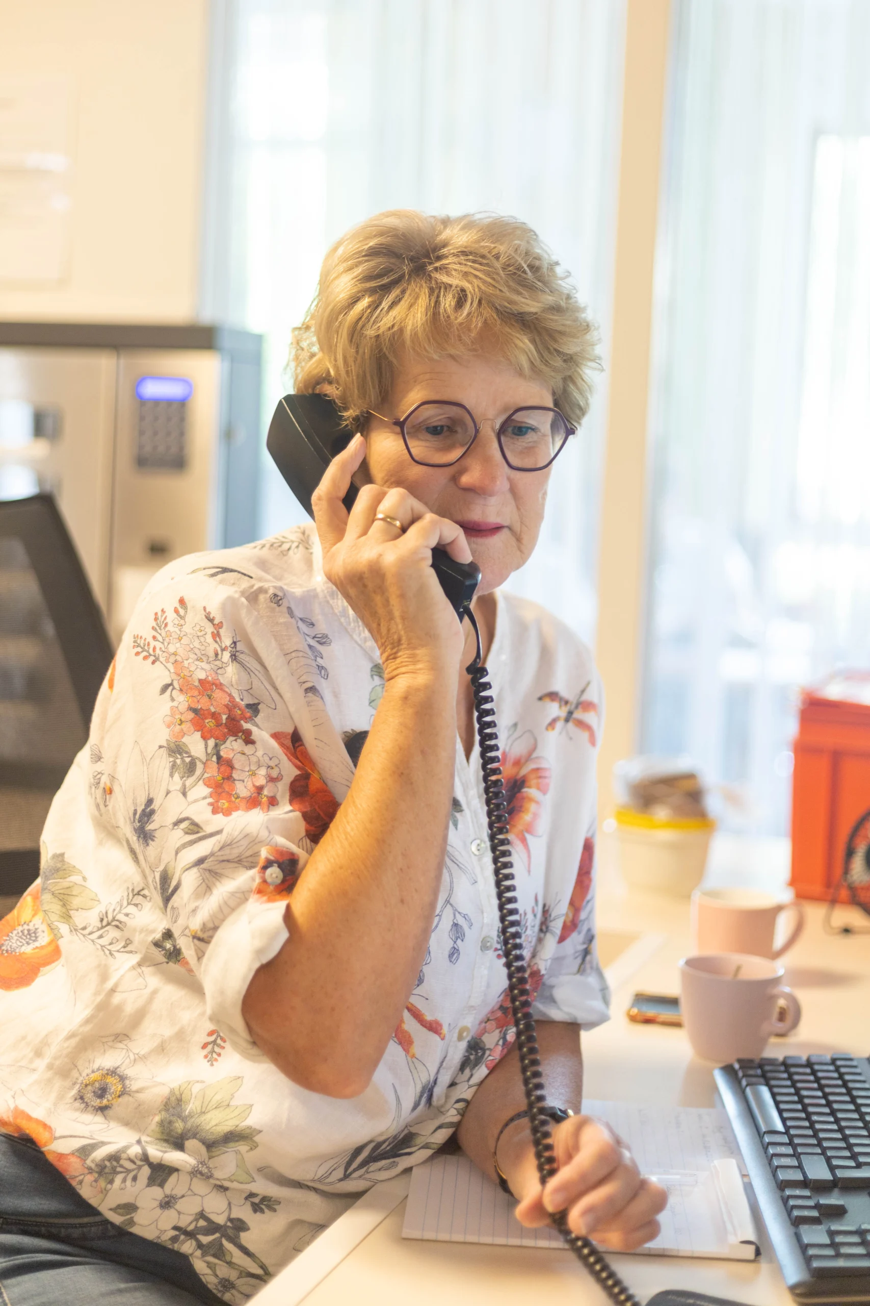 Vrouw met korte krullen en bril aan telefoon op werkplek met notitieboekje en kopjes