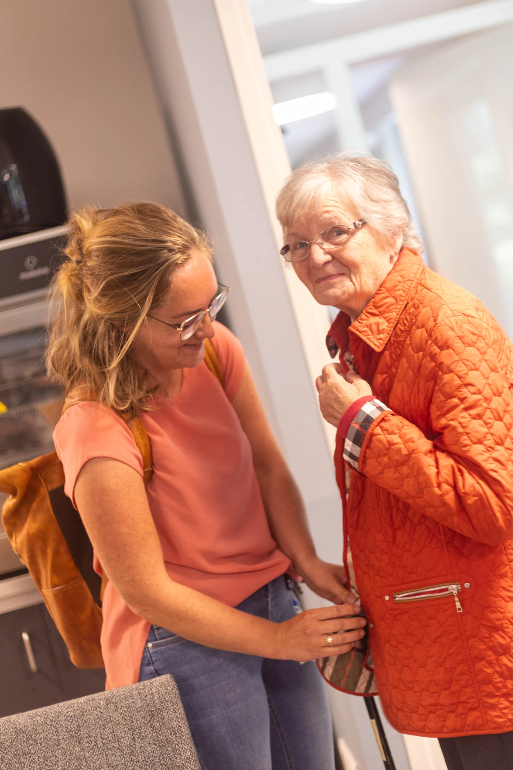 Twee vrouwen in een moderne ruimte, één draagt lichtroze shirt met rugzak, de ander oranje jas, glimlachend