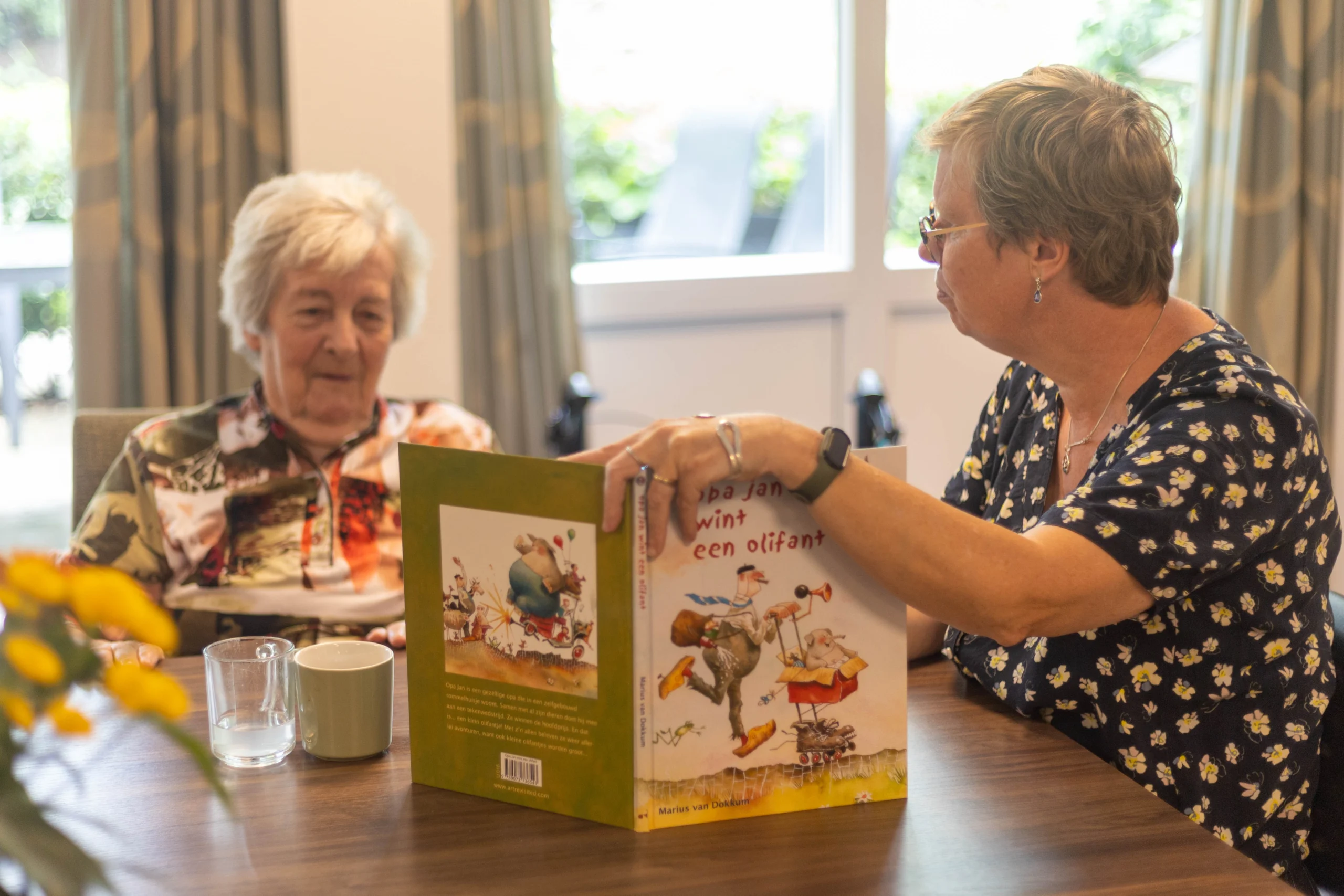 Twee vrouwen zitten samen aan tafel; een vrouw leest een boek met titel 'Opa Jan wint een olifant'.