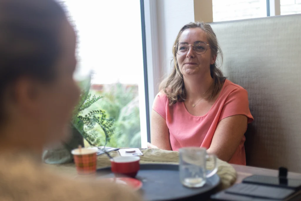 Vrouw in roze shirt met grote ronde bril aan tafel in sfeervolle ruimte met raam en groen uitzicht