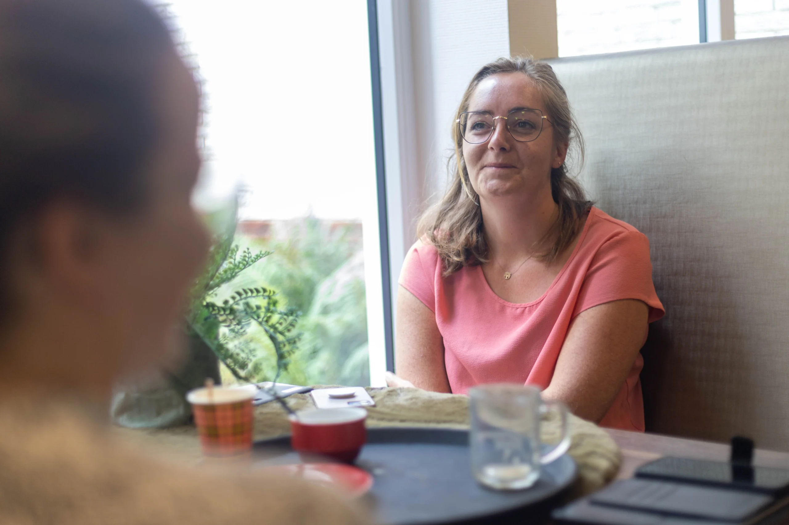 Vrouw in roze shirt met grote ronde bril aan tafel in sfeervolle ruimte met raam en groen uitzicht