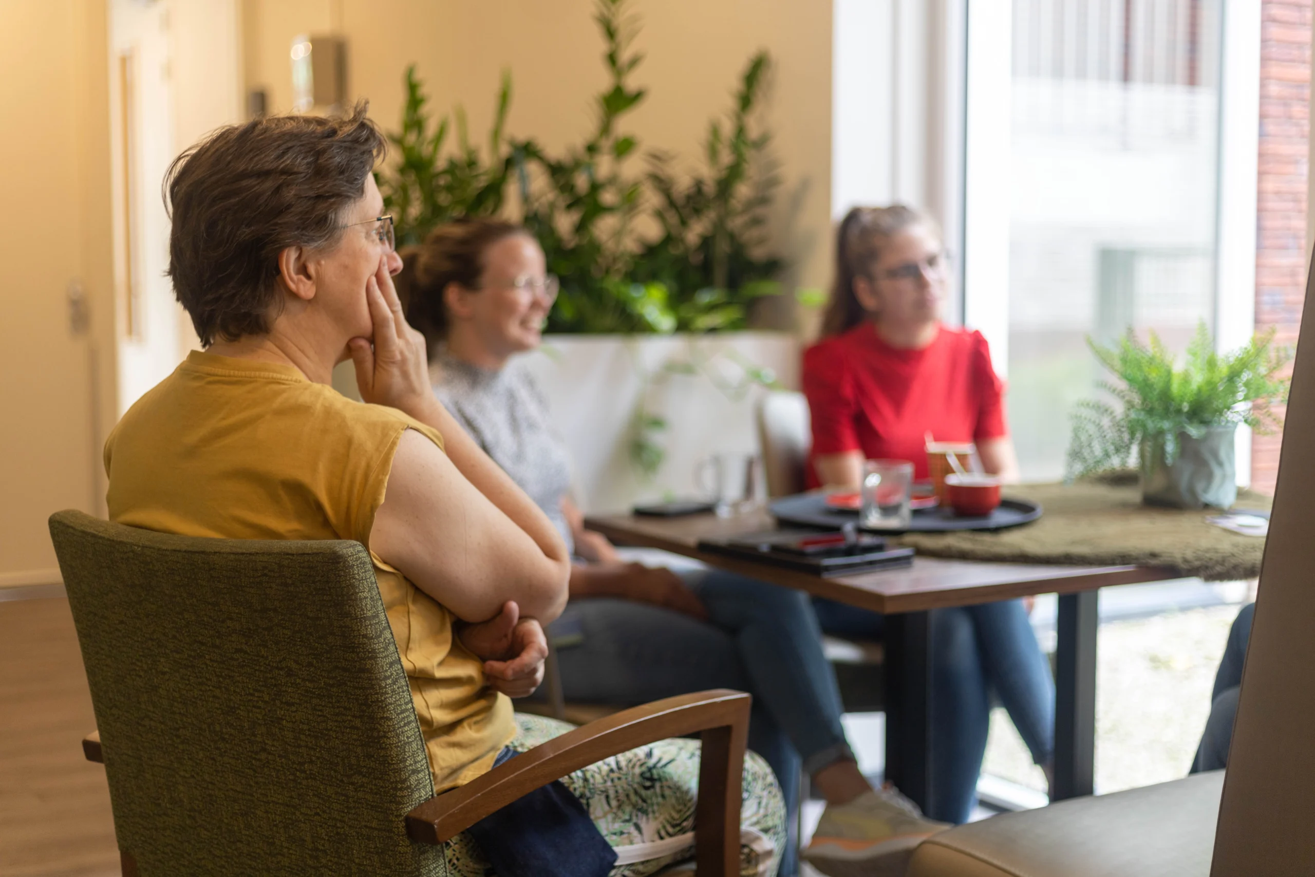 Drie vrouwen zitten samen aan een tafel met glazen en planten achter hen in een lichte ruimte.