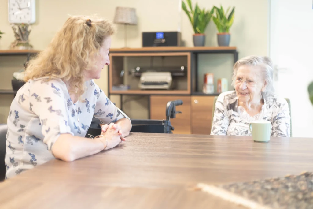 Twee vrouwen zitten aan een tafel, een jonger met krullend haar en een oudere met grijs haar en een groene mok.
