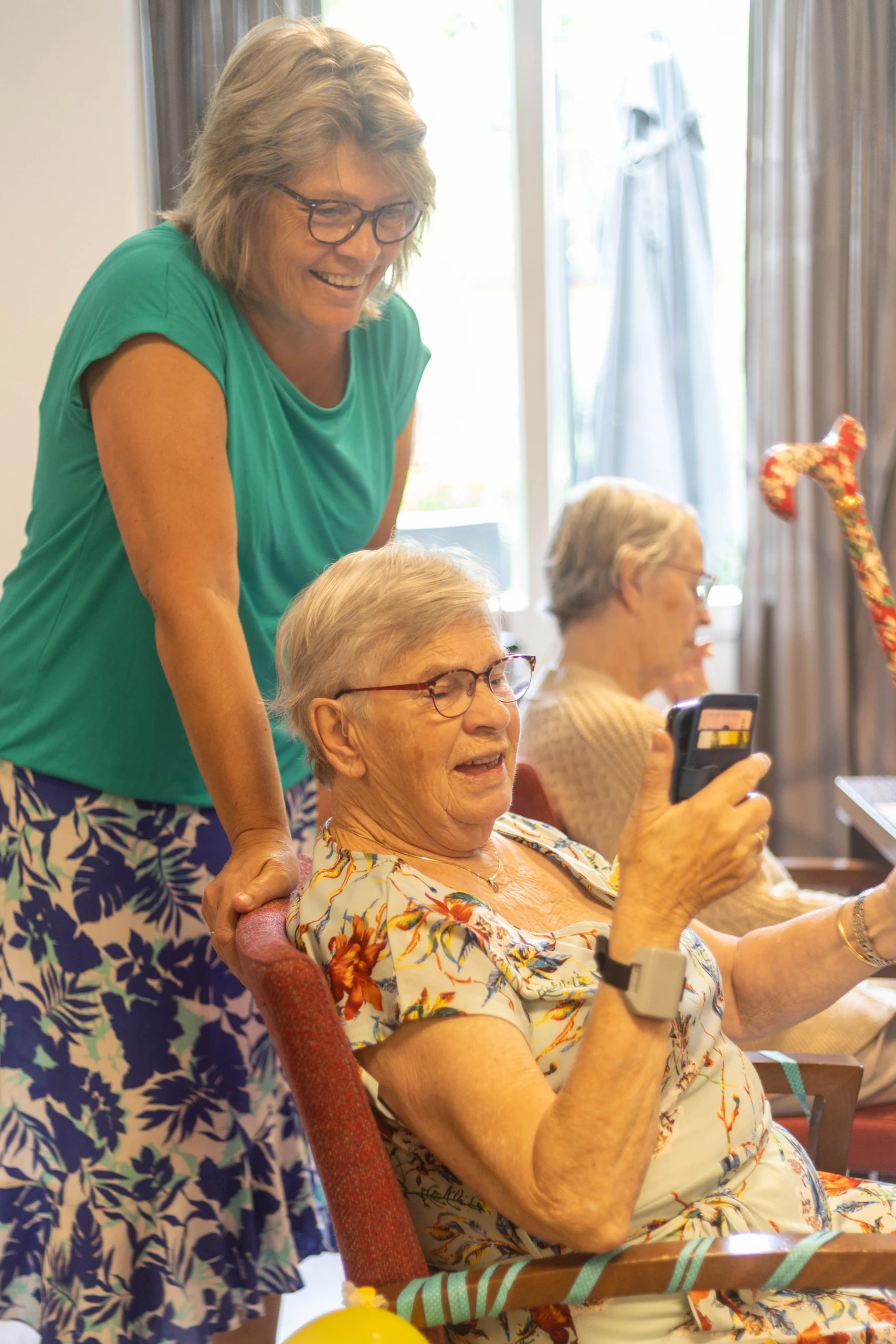 Oudere vrouw kijkt glimlachend naar apparaat in hand, jongere vrouw staat lachend achter haar
