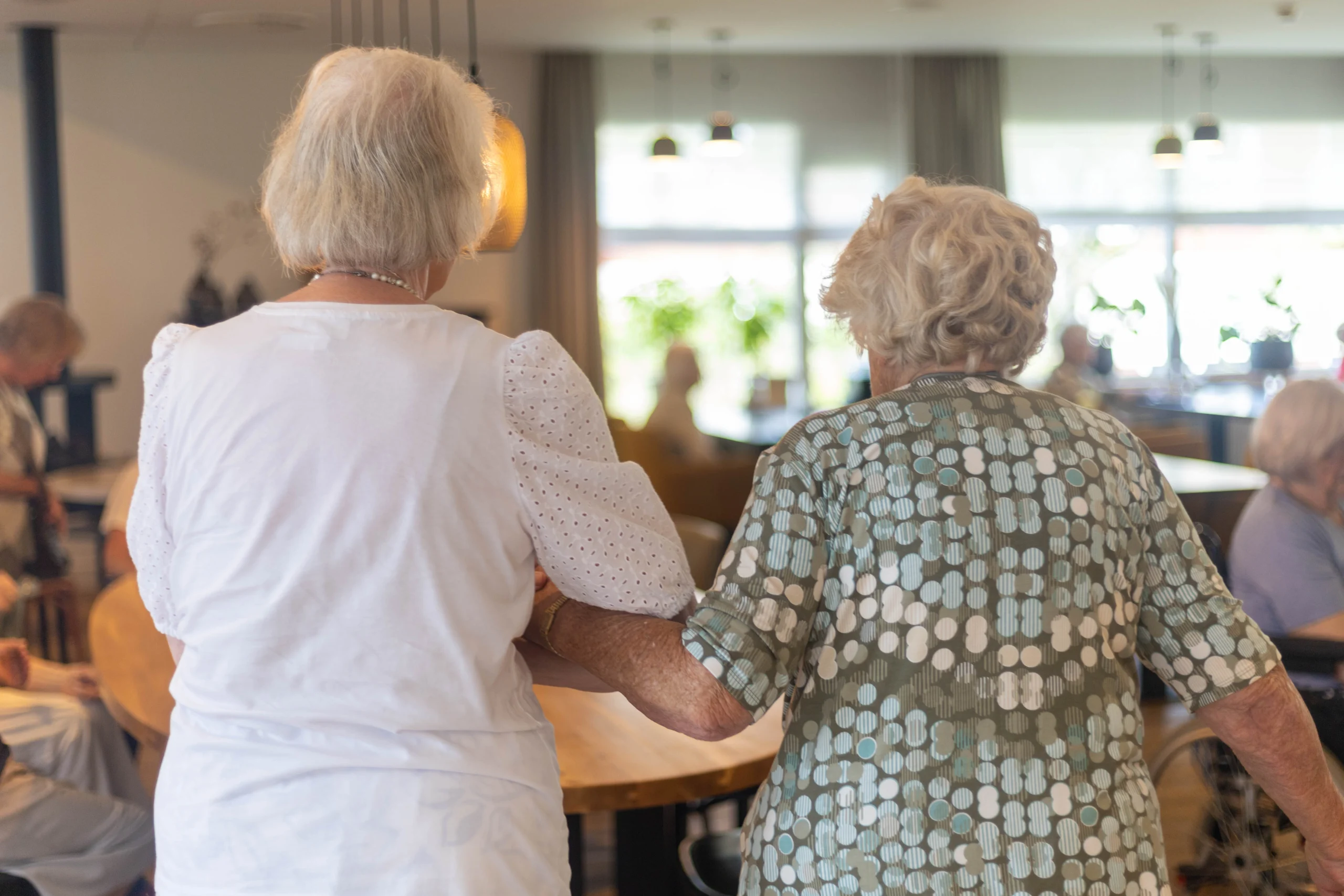 Twee oudere vrouwen lopen hand in hand in een ruimte met meerdere mensen, tafels en planten.