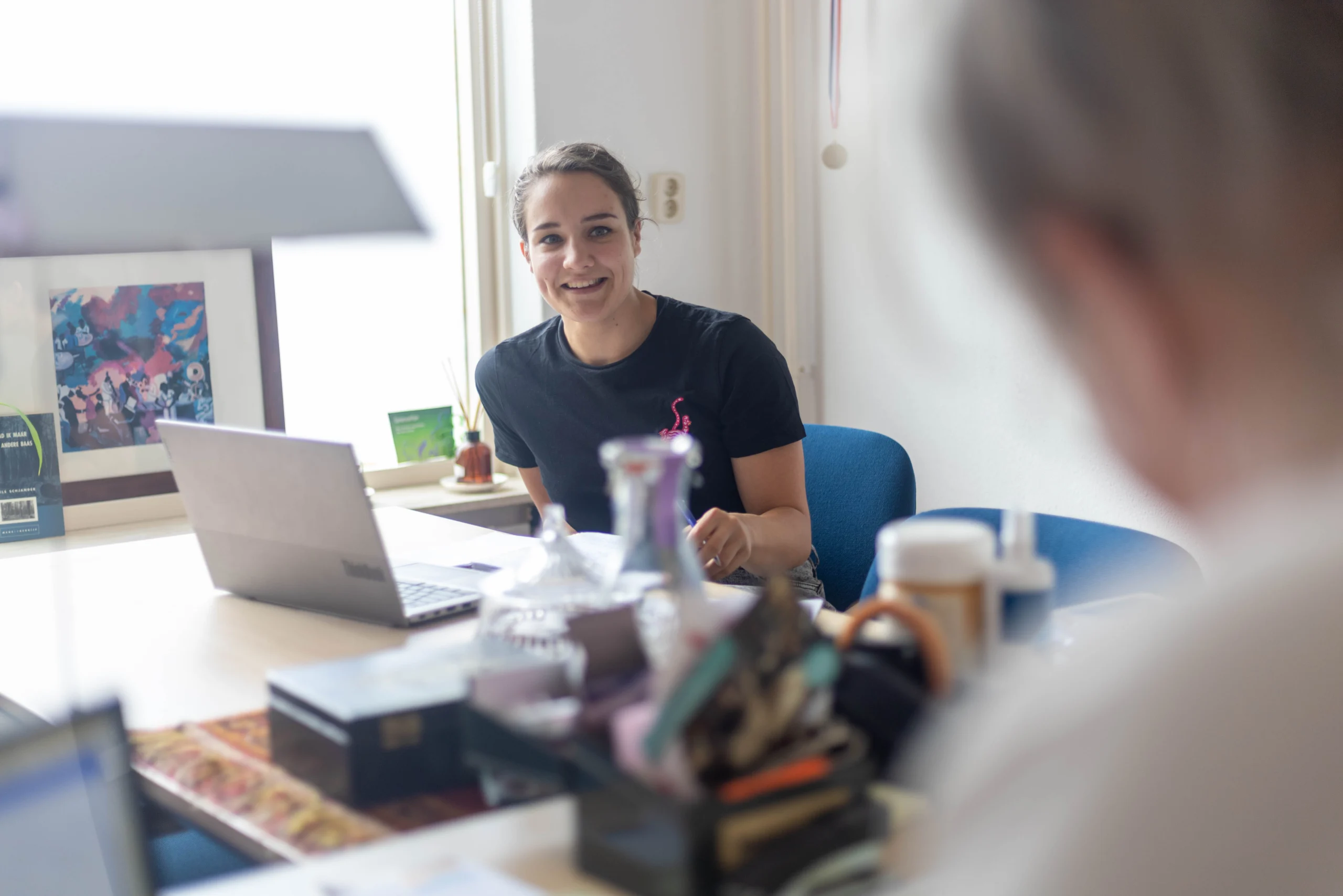 Vrouw zit aan bureau met glimlach, draagt zwart t-shirt, laptop en pennen op tafel, raam en schilderij op achtergrond