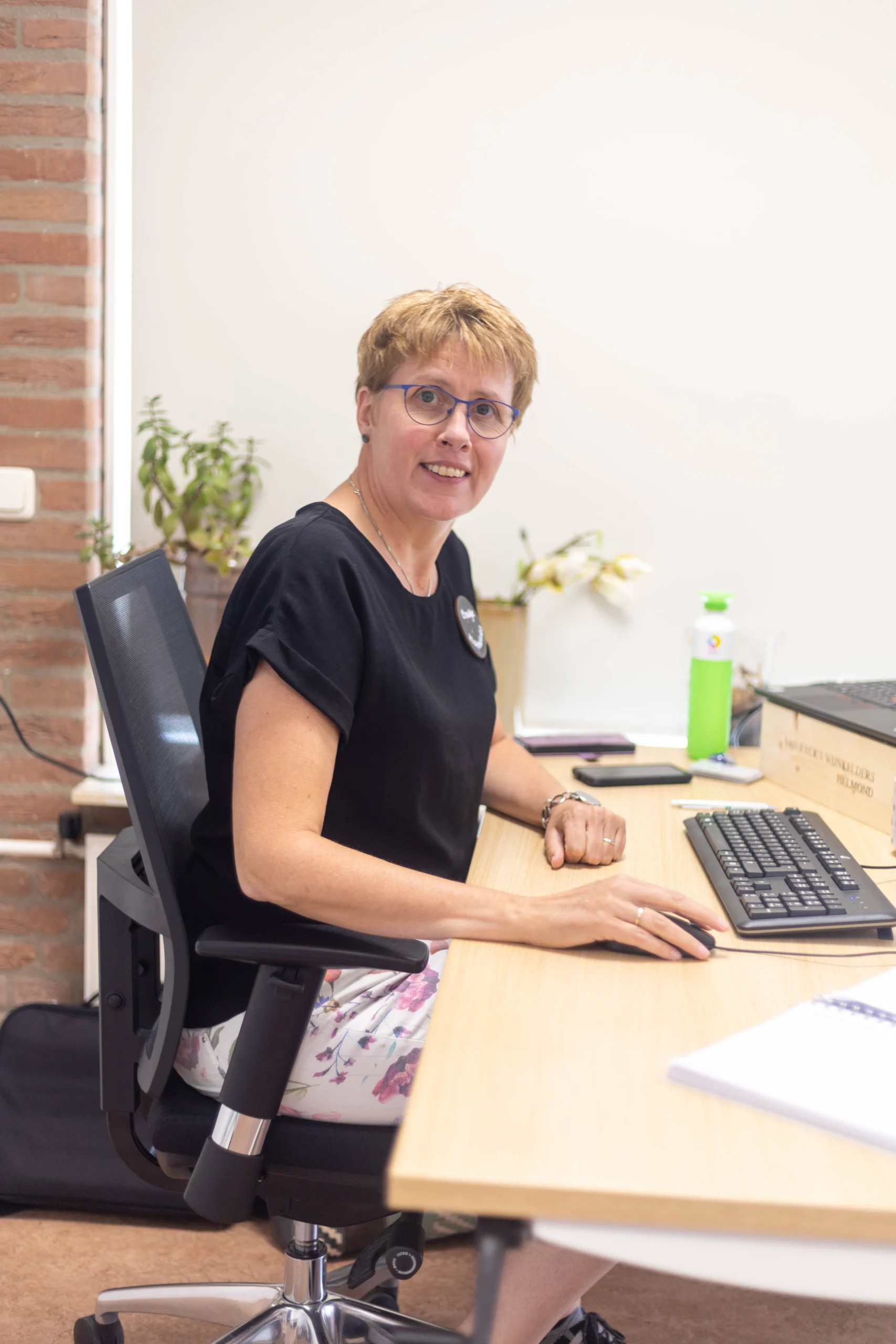 Vrouw met kort lichtbruin haar en paarse sjaal zit lachend aan een bureau met toetsenbord en planten.