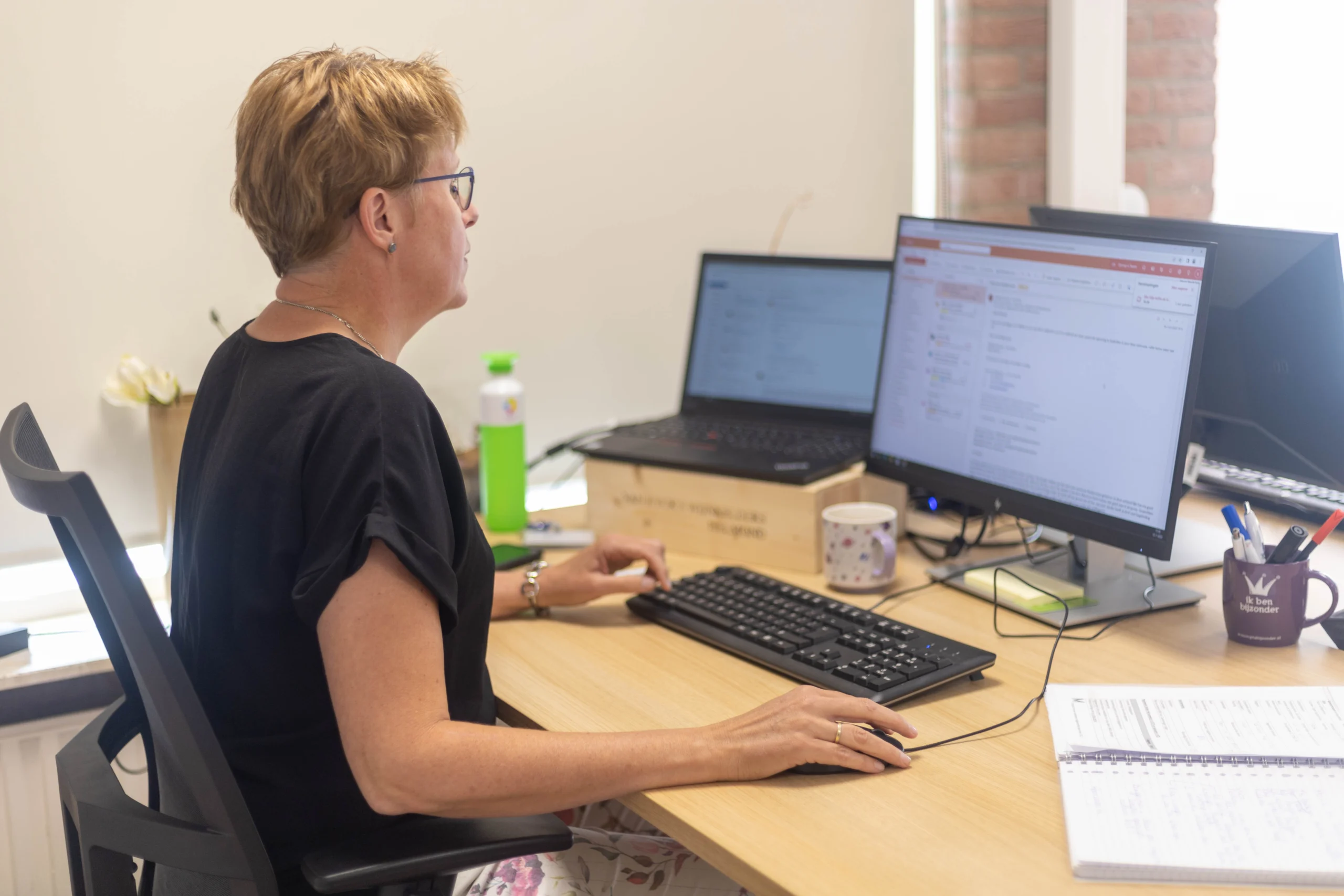 Vrouw met kort gekleurd haar zit aan bureau met laptop, twee schermen, mok en waterflesje