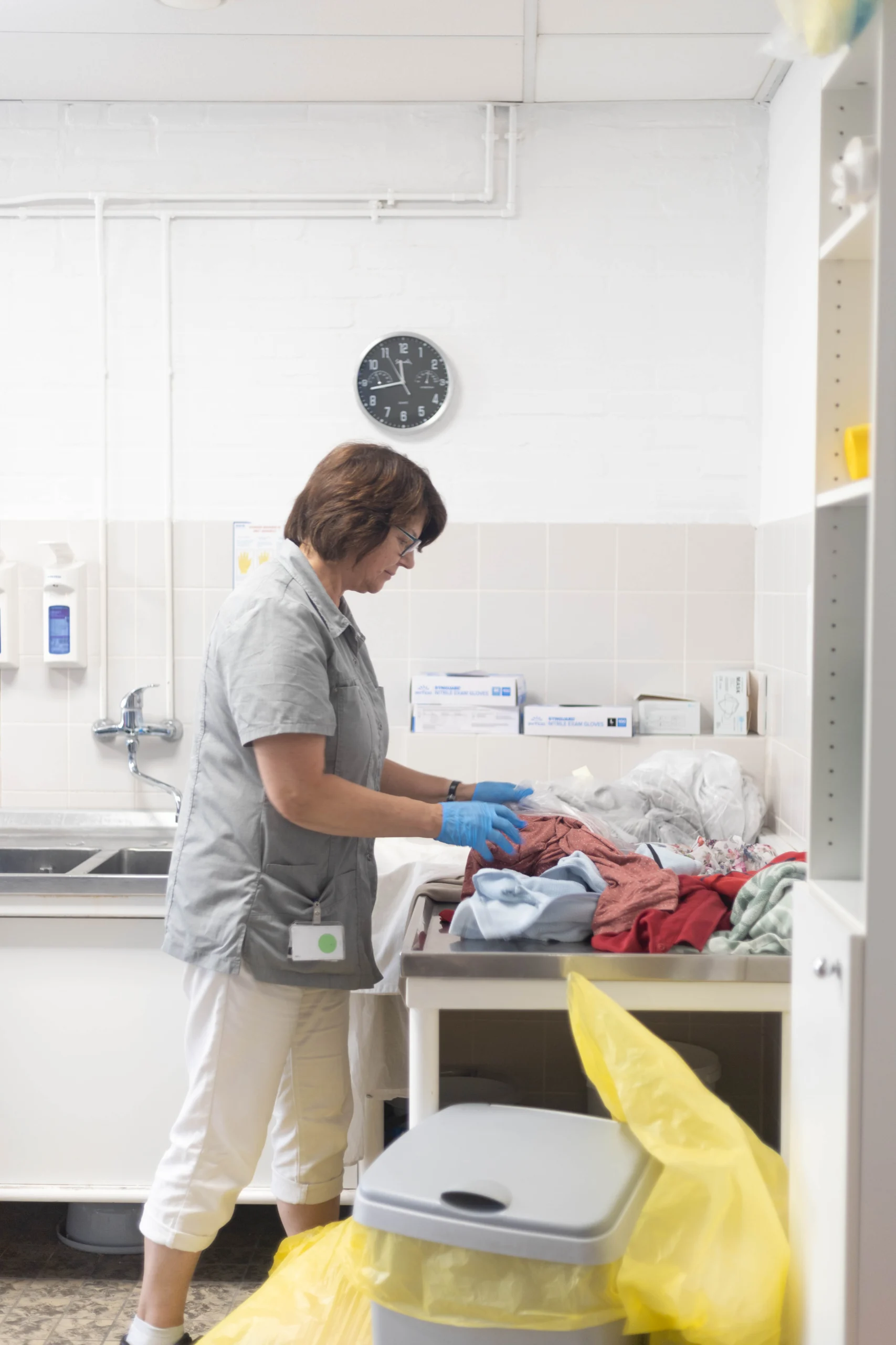 Vrouw in grijs uniform sorteert kleding in heldere wasruimte met klok en dozen