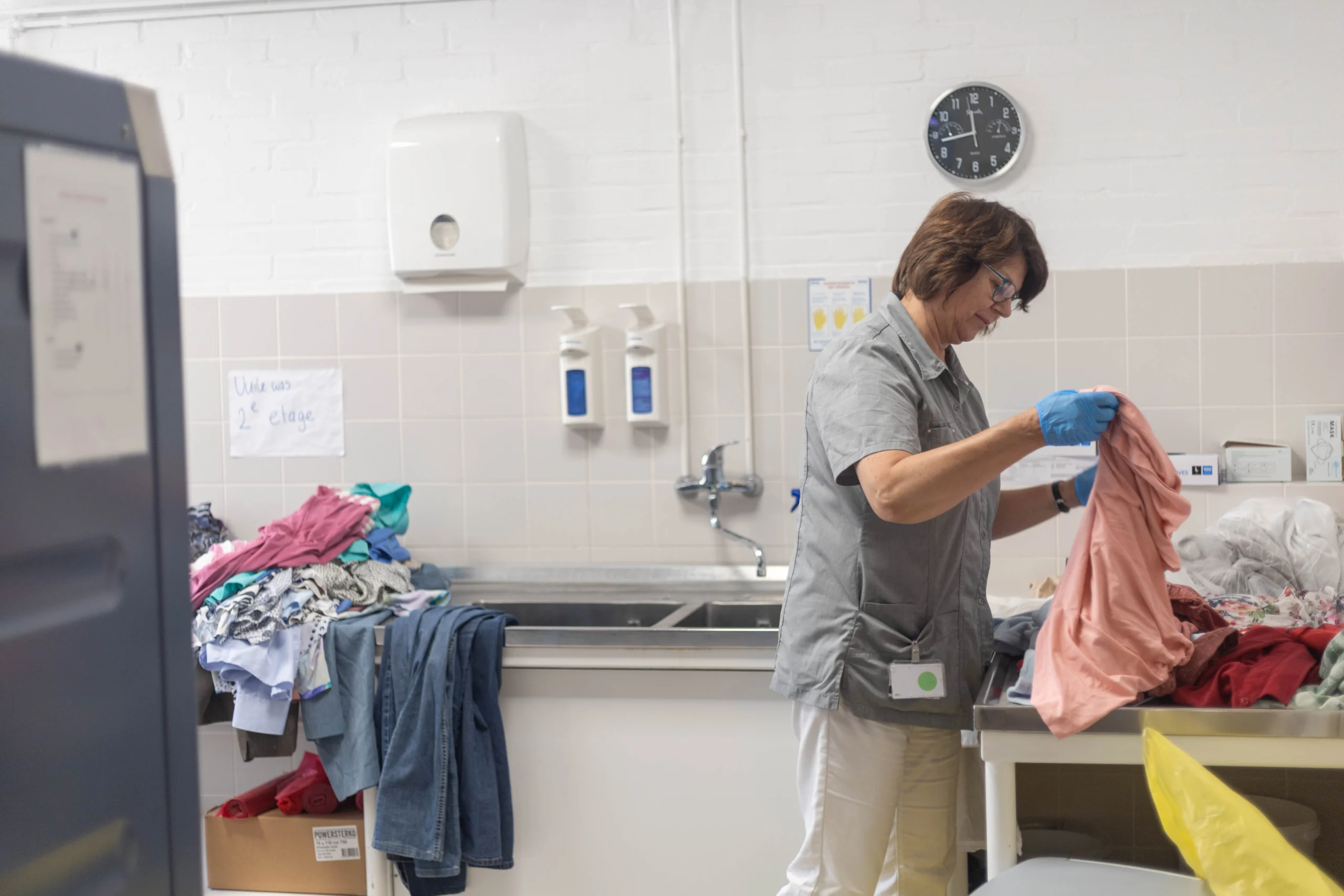 Vrouw in grijze schort en blauwe handschoenen sorteert kleding in een georganiseerde wasruimte met een gootsteen en klok.