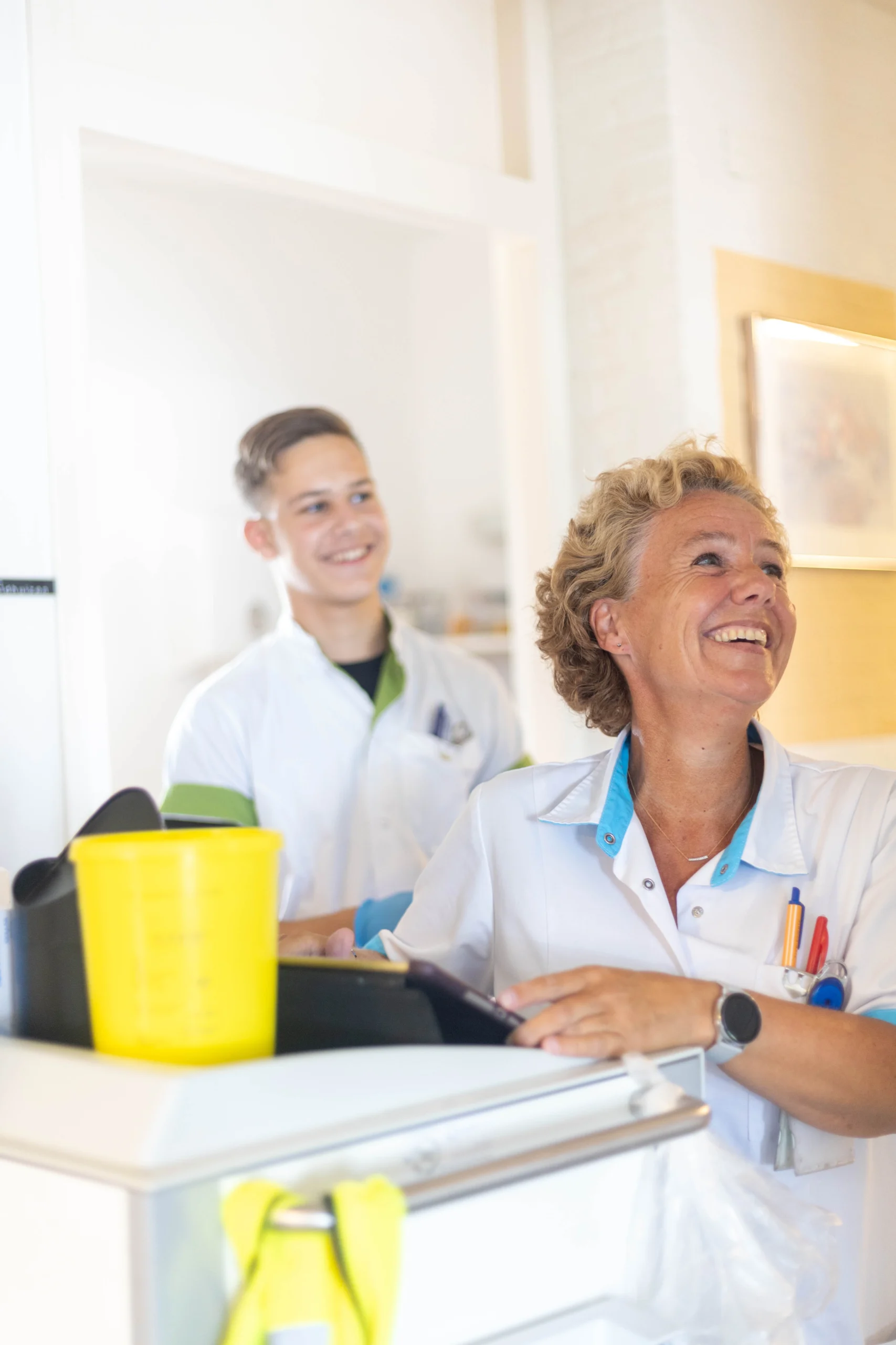 Een vrouw en een jongen in witte schorten met pennen, glimlachend in een lichte kamer met tablet en gele beker.
