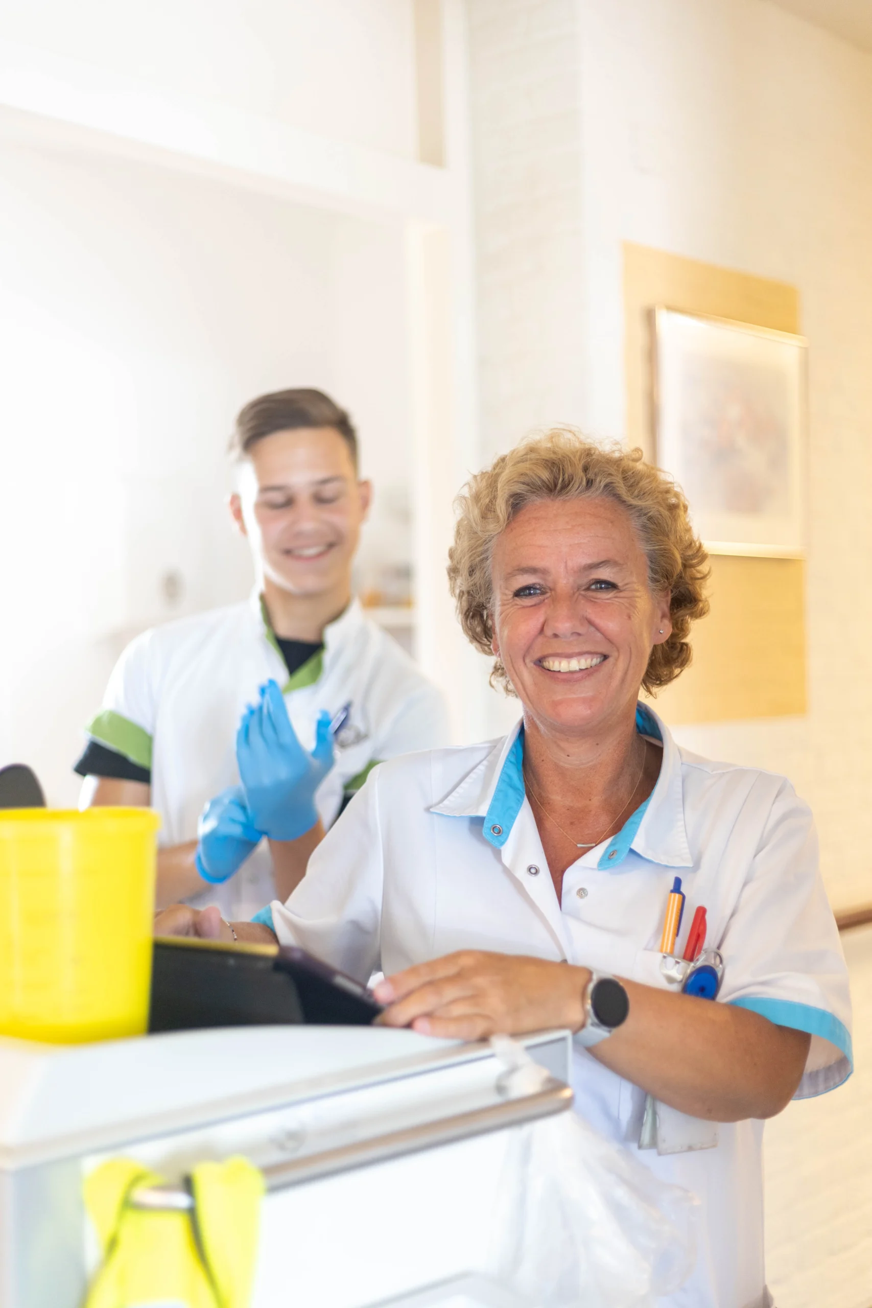 Vrouw in witte schort met tablet bij aanrecht, lachende man met witte outfit en blauwe handschoenen naast haar.