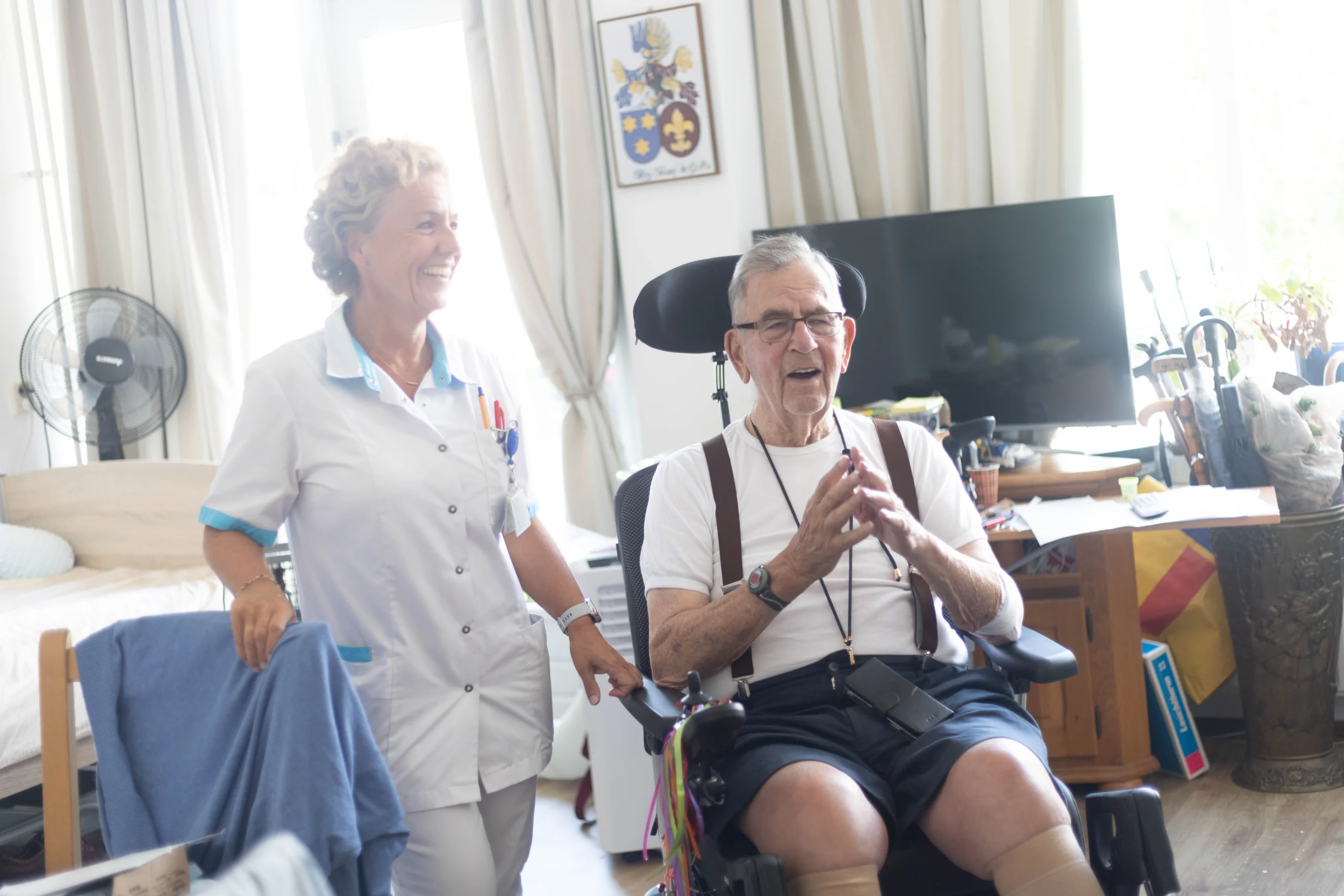 Een vrouw in verpleegkundige uniform praat lachend met een oudere man in een rolstoel in een huiselijke verpleegruimte.