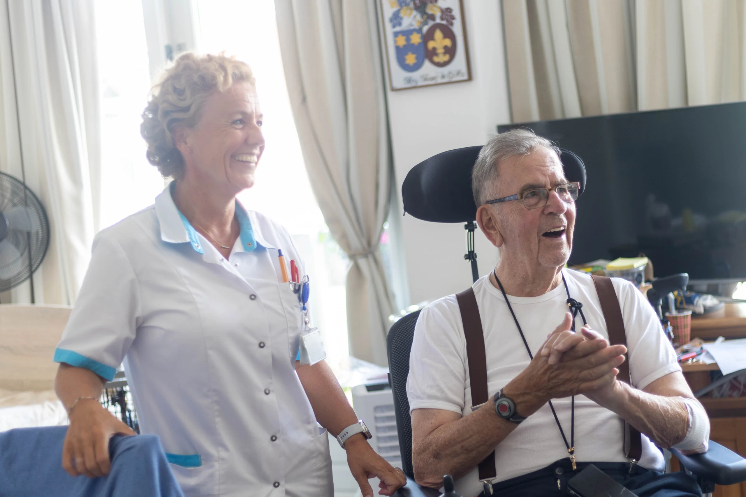 Glimlachende vrouw in verpleegsteruniform naast oudere man in rolstoel in lichte woonruimte