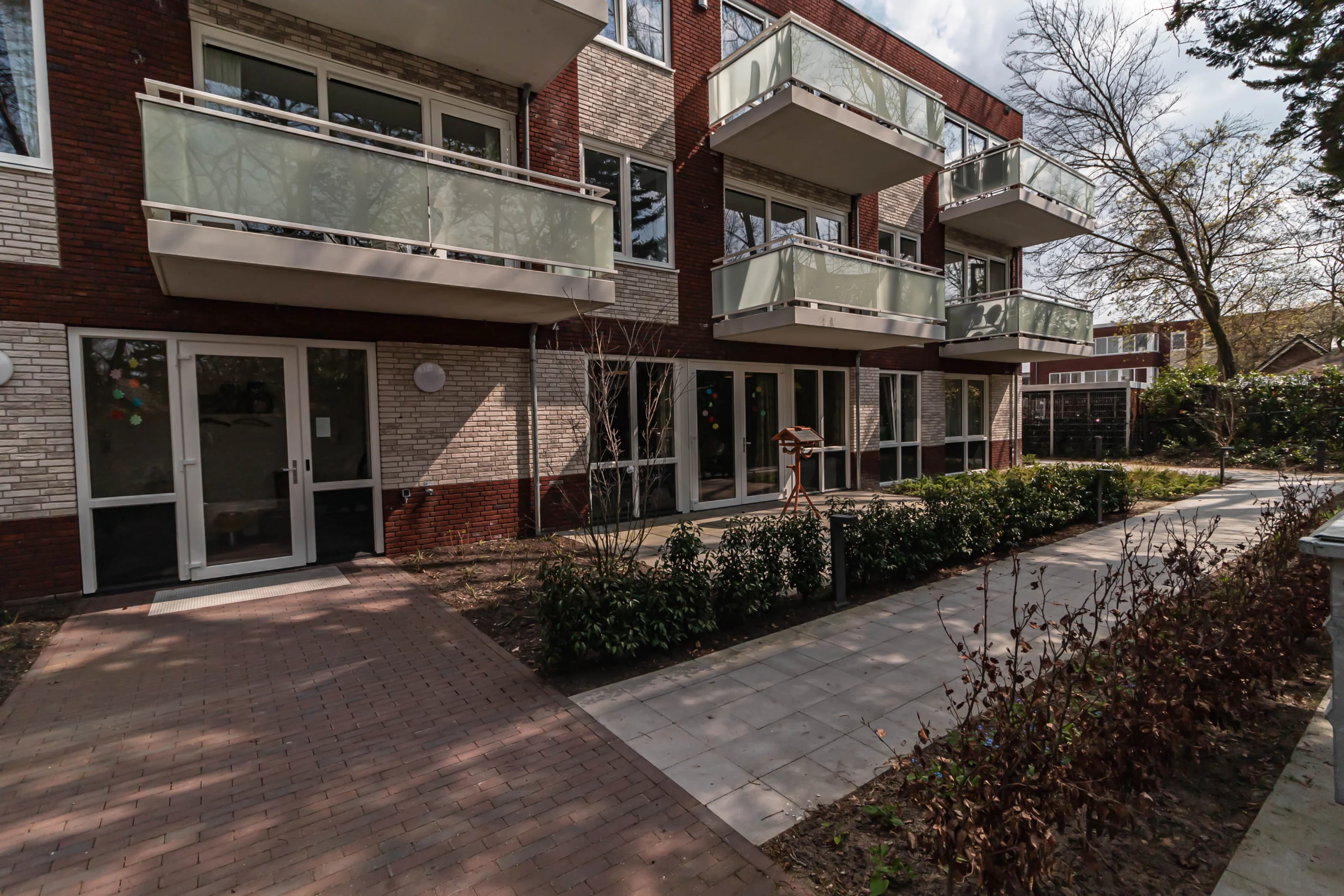 Modern appartementencomplex met rode en lichte bakstenen, balkons met glazen balustrades en groene plantenbedden.