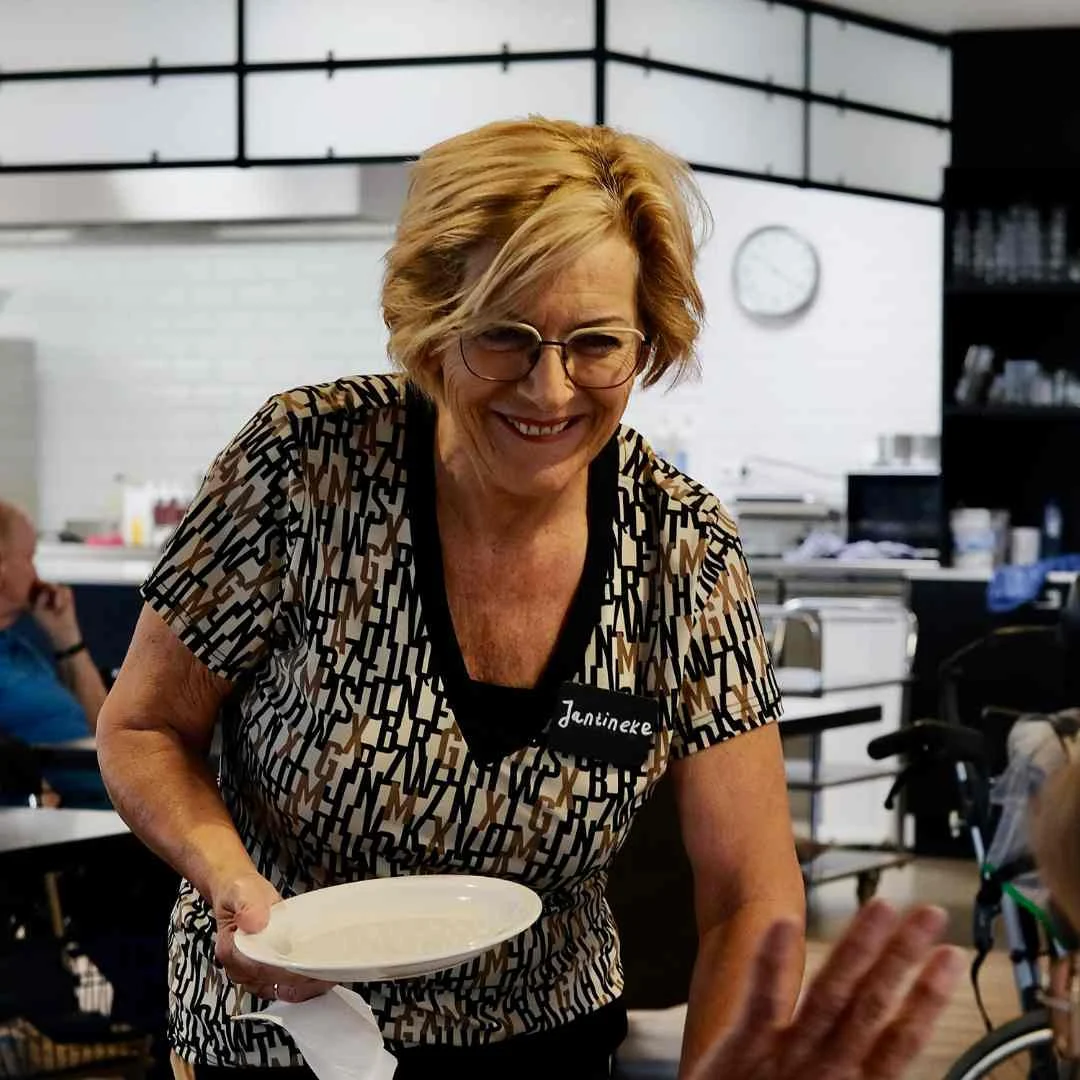 Vrouwelijke serveerster met blond haar en naamkaartje Janineke biedt een wit bord aan in eetruimte