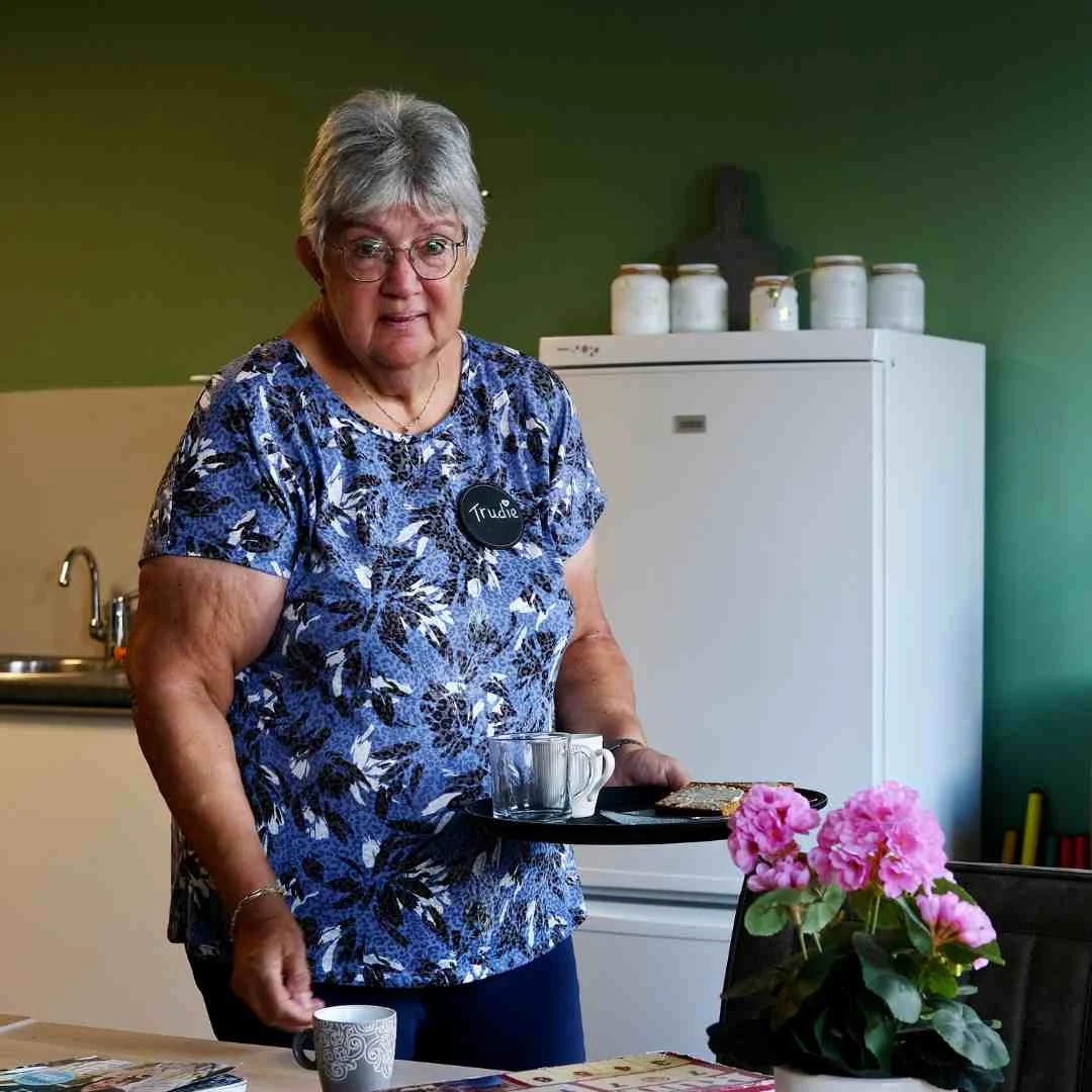 Oude vrouw met grijs haar en blauwe blouse draagt dienblad met glazen in keukenruimte