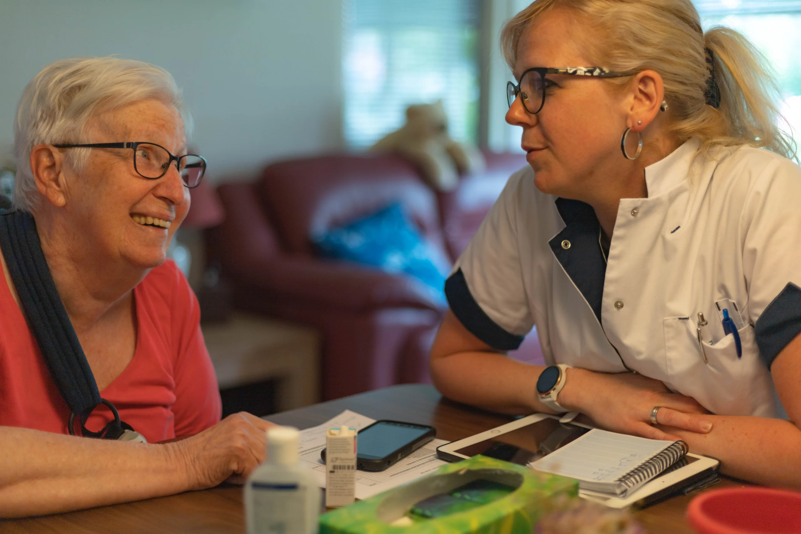 Twee vrouwen aan tafel, oudere dame met bril en roze shirt links, jongere vrouw in wit uniform rechts.