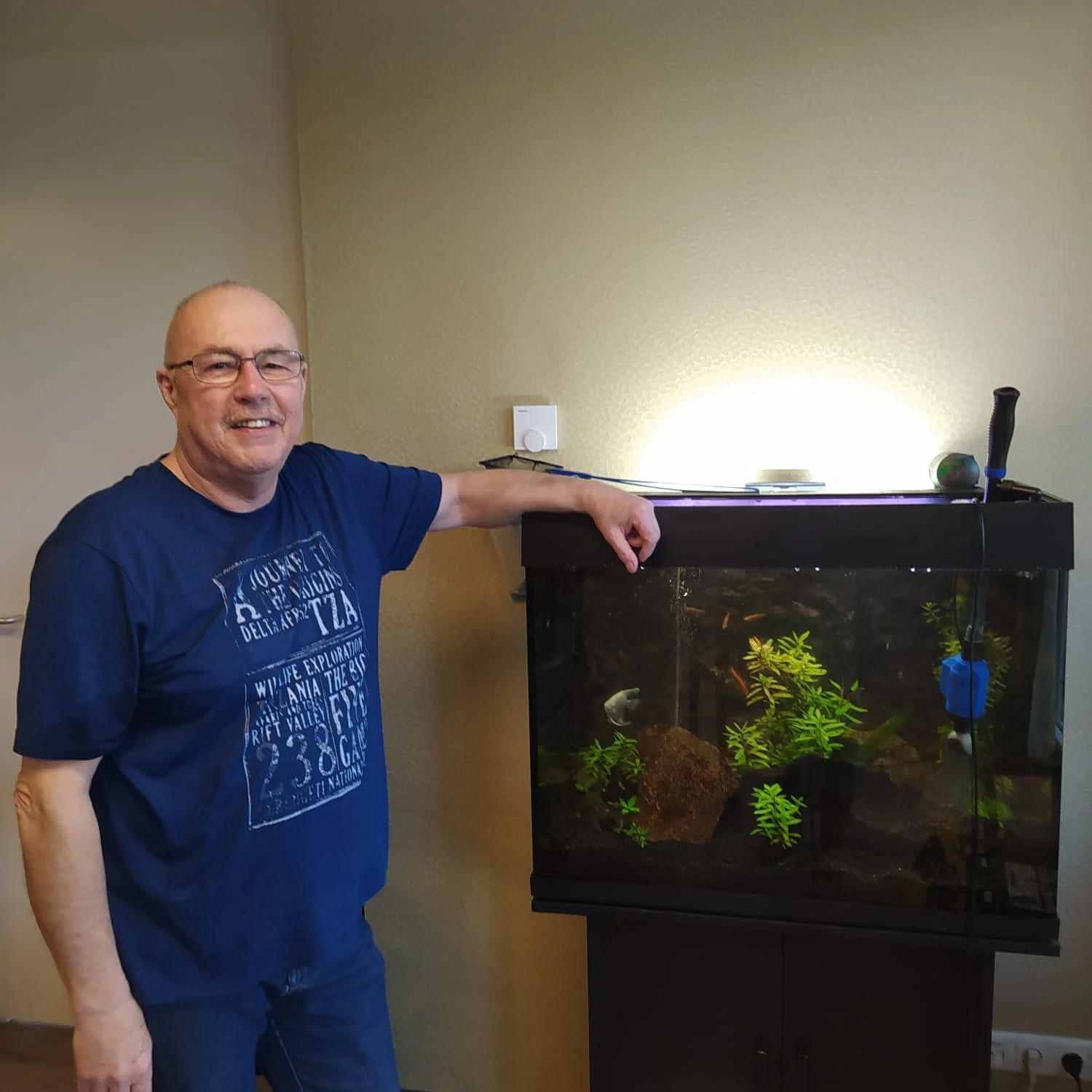 Een man met blauw T-shirt glimlacht naast een goed verlicht aquarium met planten en vissen.