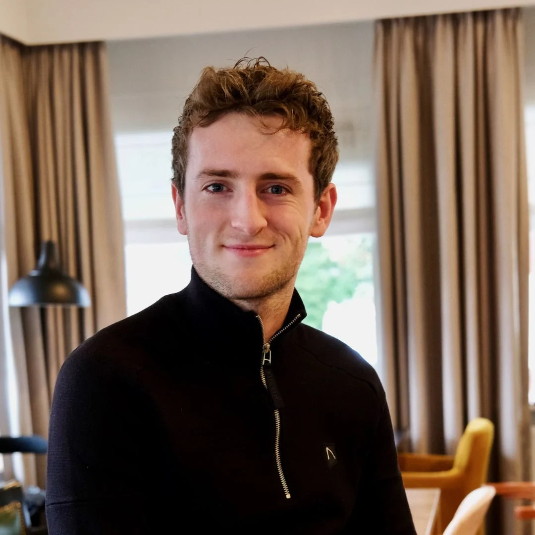 Jongeman met zwarte trui en hoge kraag glimlacht in lichte binnenruimte met raam en gordijnen