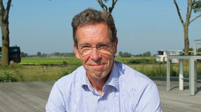 Man in lichtblauw gestreept overhemd met bril glimlacht zittend op houten terras met bomen en groene velden op achtergrond