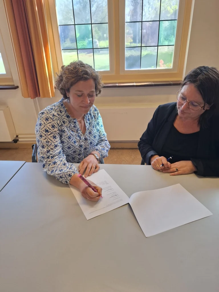 Twee vrouwen zitten aan een tafel, een schrijft met roze pen, de ander kijkt naar een document.