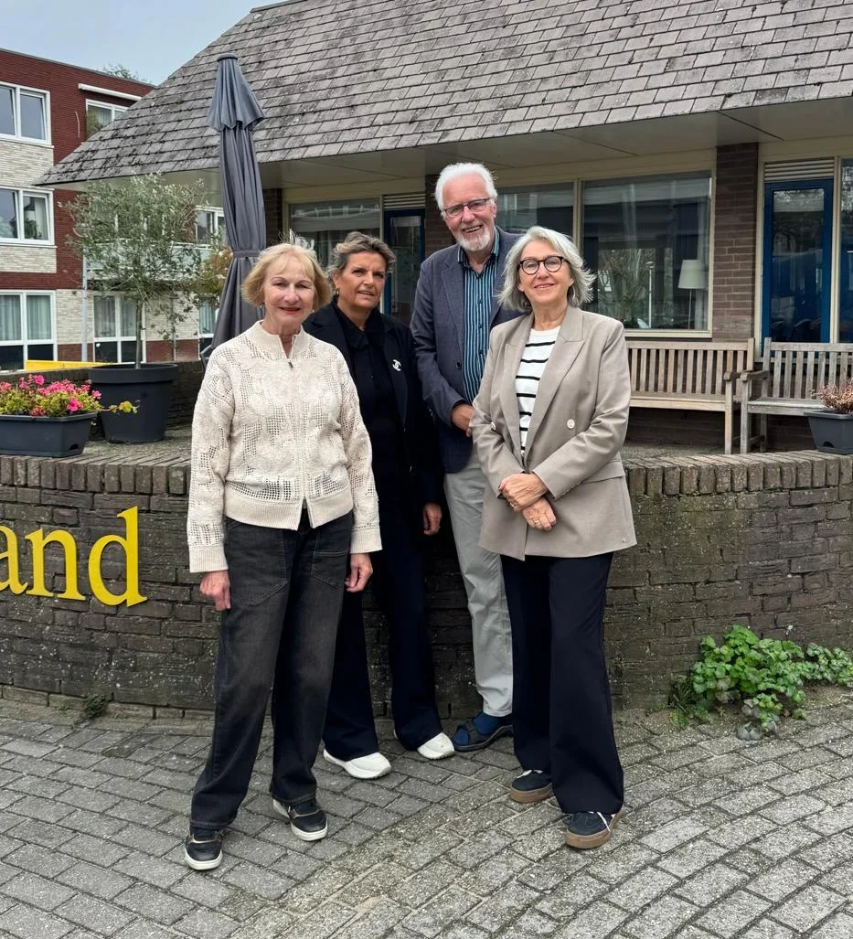 Vier mensen staan buiten voor een gebouw met laag puntig dak en muurtekst 'Onderzand'.
